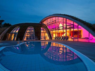 Modernes Hotel mit beleuchtetem Glasdach und Pool bei Dämmerung.