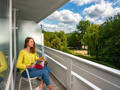 Frau sitzt auf Balkon mit Buch, umgeben von grünem Park und blauem Himmel.