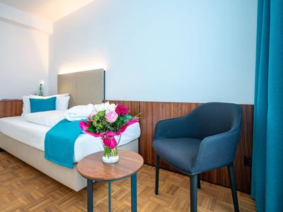 Schlichtes Hotelzimmer mit Einzelbett, blauem Bettläufer, Stuhl und Blumen auf kleinem Tisch.