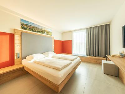 Camera d'hotel luminosa con letto matrimoniale, ampia finestra e TV a muro.