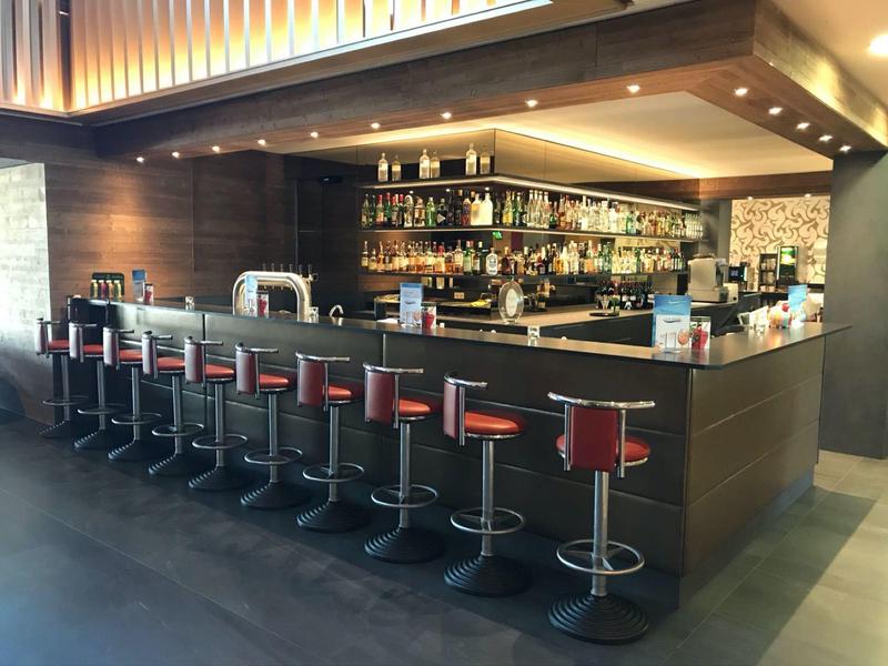 Bar d'hotel moderna con un lungo bancone e diversi sgabelli rossi.