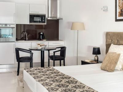 Modernly furnished hotel room with double bed, dining table, and kitchenette in neutral colors.