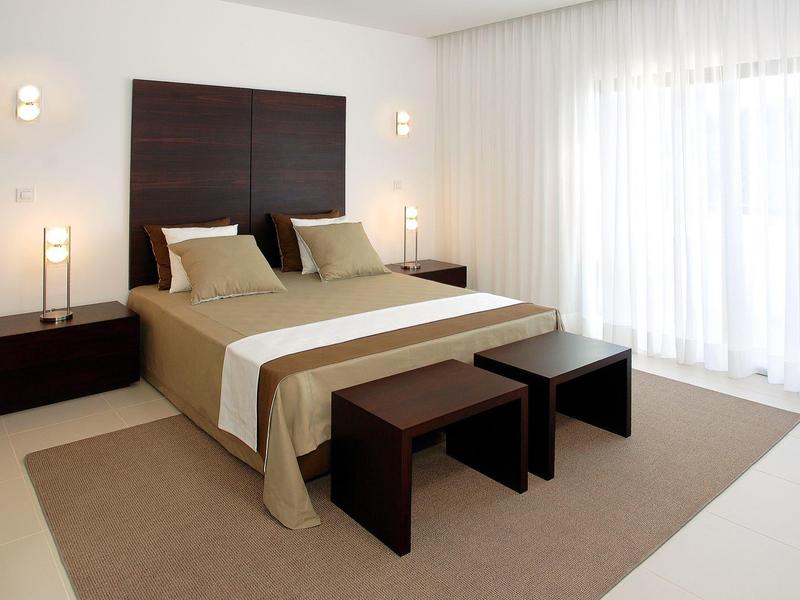 Modern hotel room with large bed, dark wooden furniture, and floor-to-ceiling white curtains.