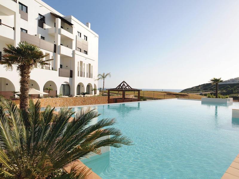 Modern hotel pool with palm trees and view of hills under clear sky.