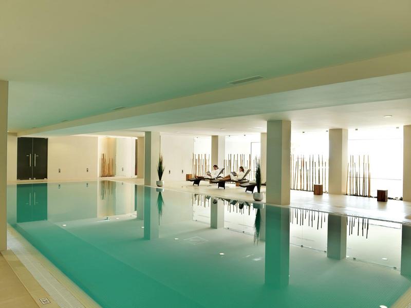Modern indoor pool with calm water and seating area with lounge chairs in the background.