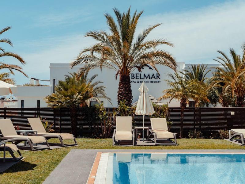 Pool with sun loungers and palm trees in front of a white building under clear sky.