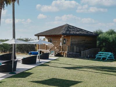 Ein kleiner Holzpavillon mit Schindeldach, umgeben von grüner Wiese, Palmen und Sitzgelegenheiten.