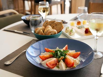 Ein Teller mit Tomatensalat und Käse, daneben Brot und Gläser mit weißem Wein auf einem Tisch.