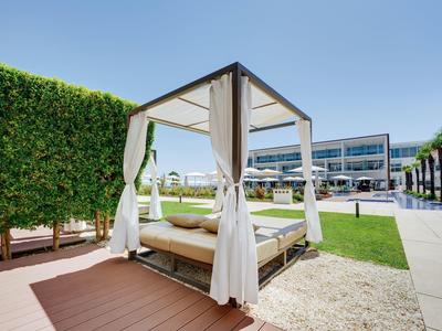 Moderner Pavillon mit weißem Stoffdach, Vorhängen und Liege auf Holzterrasse neben grüner Hecke.