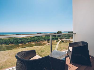 Balkon mit zwei schwarzen Stühlen, Gläsern und Ausblick auf grüne Wiese, Meer und blauen Himmel.