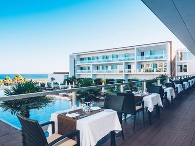 Blick auf Hotelterrasse mit Tischreihen am Pool und Meer im Hintergrund unter blauem Himmel.