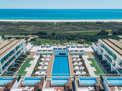 Symmetrisches Hotel mit blauem Pool, Liegestühlen und direktem Zugang zum Strand und Meer.