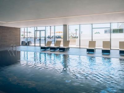 Innenraum mit großem rechteckigem Pool, großen Fenstern und mehreren weißen Liegestühlen.