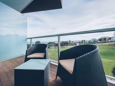 Moderner Balkon mit Glasgeländer, schwarzem Rattansessel, Hocker und grünem Rasen im Hintergrund.