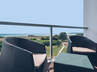 Balkon mit zwei schwarzen Geflechtstühlen, blauem Tisch und Blick auf grünes Feld und Meer im Hintergrund.