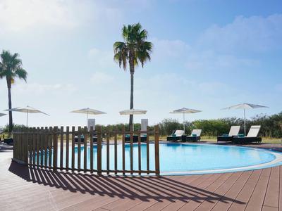 Runder Pool mit Holzdeck, Liegestühlen, Sonnenschirmen und Palmen unter blauem Himmel.