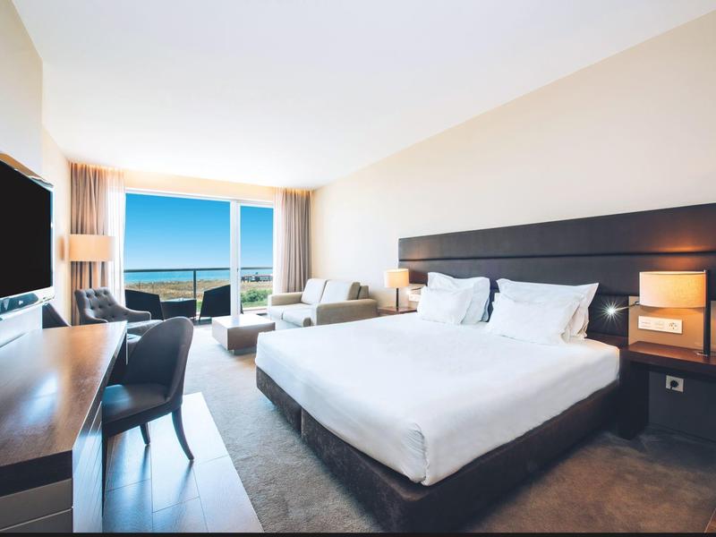 Jasny pokój hotelowy z łóżkiem podwójnym, miejscem do siedzenia, biurkiem i widokiem na morze przez duże okna.