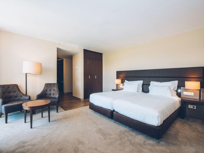 Nowoczesny pokój hotelowy z podwójnym łóżkiem, dwoma fotelami i małym okrągłym stolikiem.