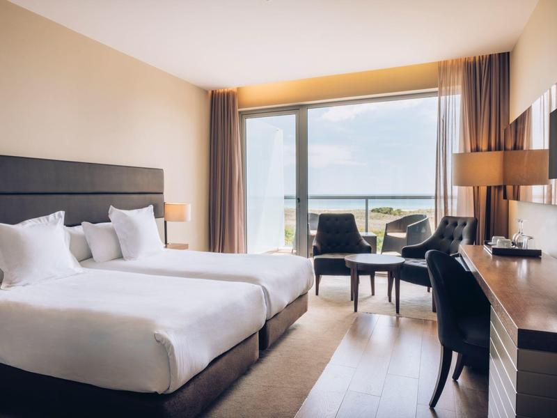 Nowoczesny pokój hotelowy z dwoma łóżkami, biurkiem i widokiem na morze przez duże okna.