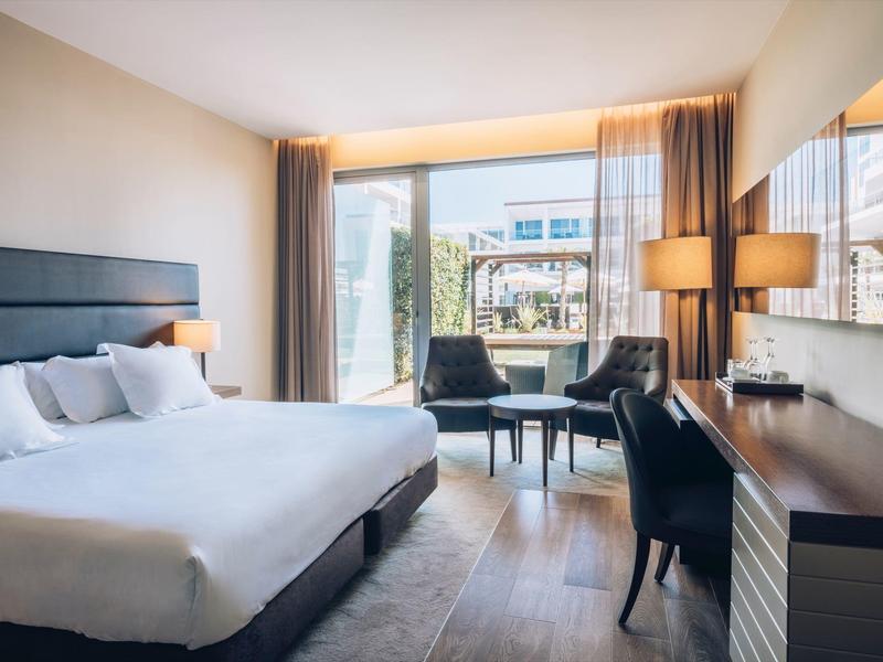 Nowoczesny pokój hotelowy z łóżkiem dwuosobowym, biurkiem, fotelami i dużym oknem z widokiem na miasto.