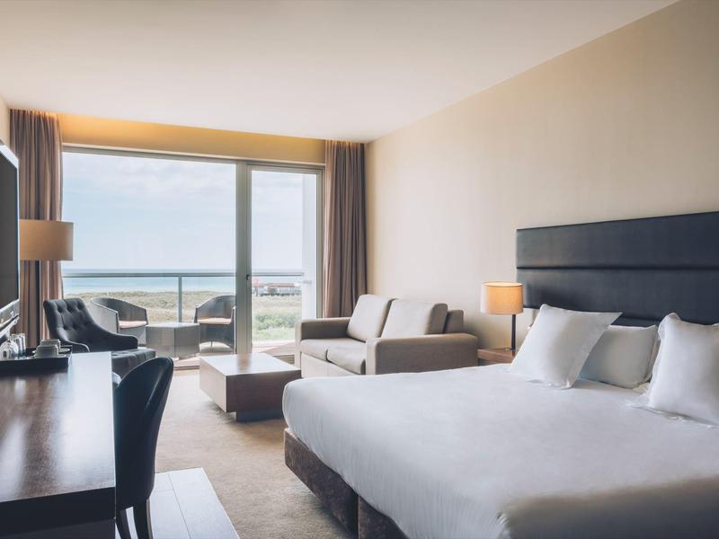 Jasny pokój hotelowy z podwójnym łóżkiem, sofą i balkonem z widokiem na morze.