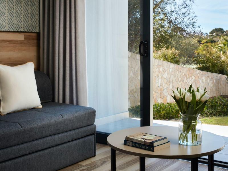 Gezellige zithoek met bank, kussen, salontafel, boeken en bloemen bij groot raam met uitzicht op tuin.