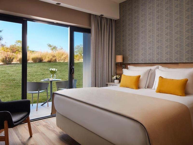 Moderne hotelkamer met groot bed, zithoek en terras met uitzicht op een groen gazon.
