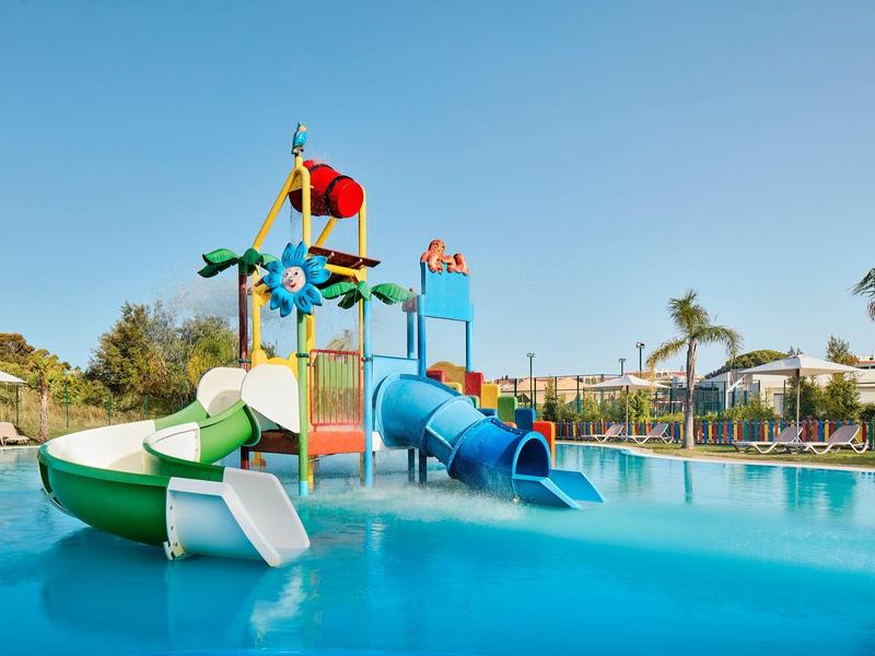 Water speelplaats met glijbanen en sproeiers in een zwembad onder een blauwe lucht.