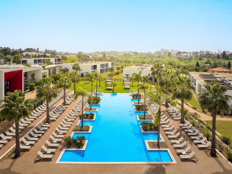 Groot rechthoekig zwembad tussen rijen ligstoelen bij een hotel met palmbomen