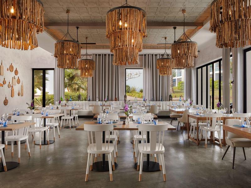 Lichte, moderne eetzaal met houten meubels, grote ramen en opvallende kroonluchters.