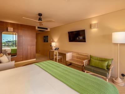 Helles Hotelzimmer mit großem Bett, grüner Decke, Sofa, Fernseher und Arbeitsbereich.