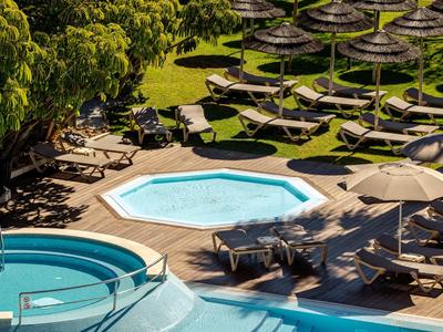 Mehrere Liegestühle und Sonnenschirme um einen kleinen Pool in einem Hotelgarten bei Tageslicht.