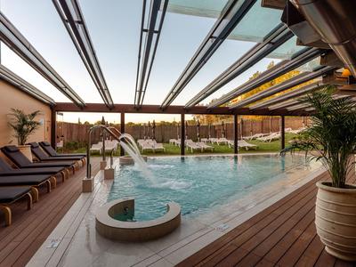 Überdachter Hotelpool mit Liegestühlen und Wasserfall, umgeben von Holzterrasse und Pflanzen.
