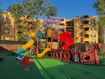 Bunter Kinderspielplatz mit Rutschen und Piratenschiff vor einem Hotelgebäude bei sonnigem Wetter.