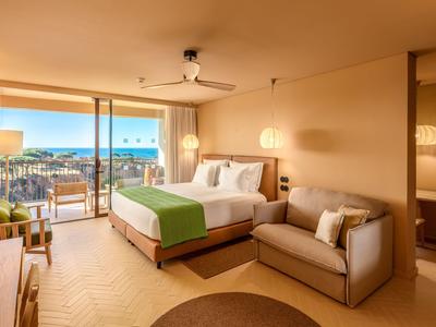 Helles Hotelzimmer mit Meerblick, großem Bett und Sitzbereich mit Sofa.