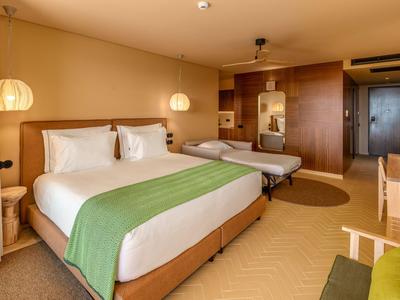 Hotelzimmer mit Doppelbett, Einzelbett, warmen Holztönen und modernen Lampen.