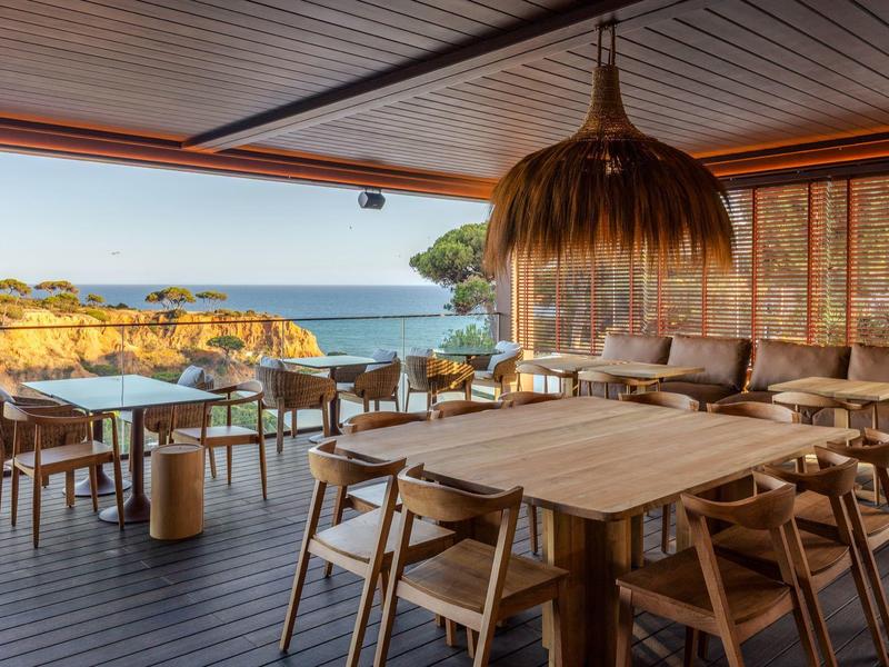 Espace de restaurant ouvert en bord de mer avec tables en bois, chaises et vue sur les falaises.