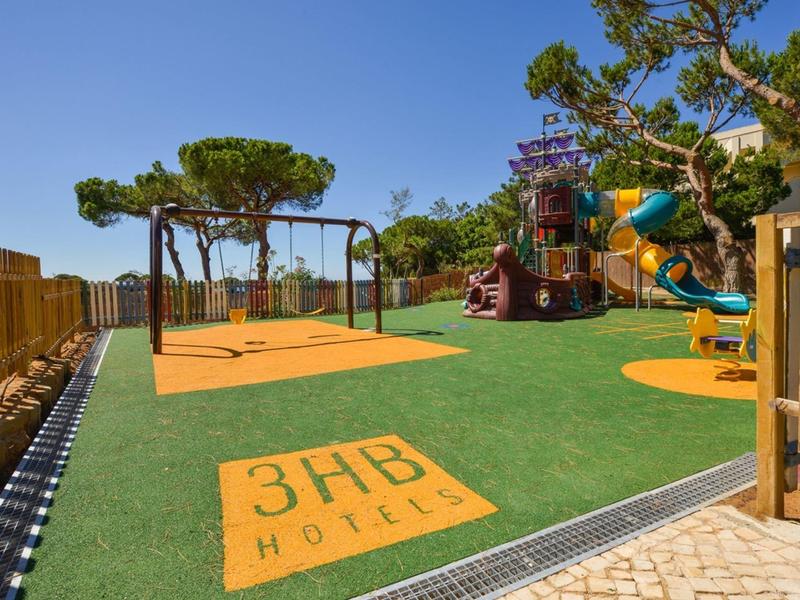 Aire de jeux avec toboggans et balançoires sur sol vert sous un ciel bleu à côté d'une clôture.