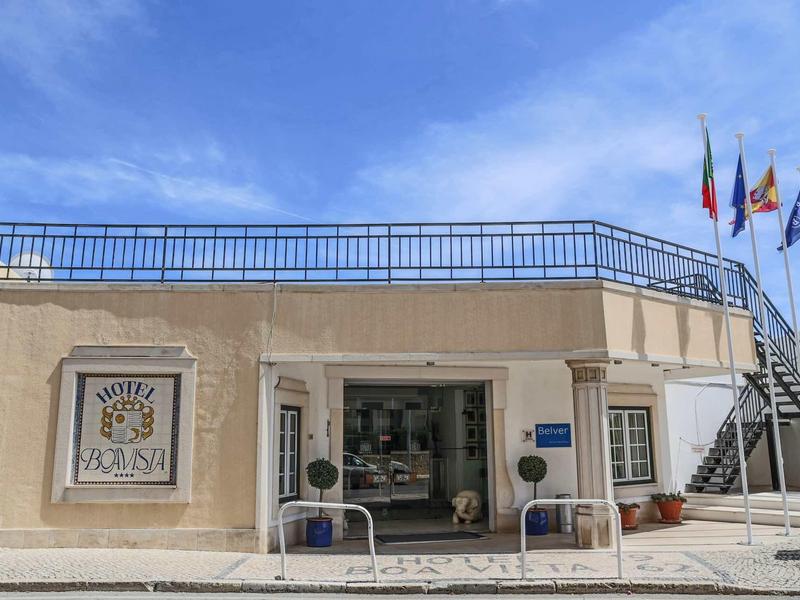 Jasna elewacja hotelu z zewnętrznymi schodami, trzema flagami i czystym niebem w tle.