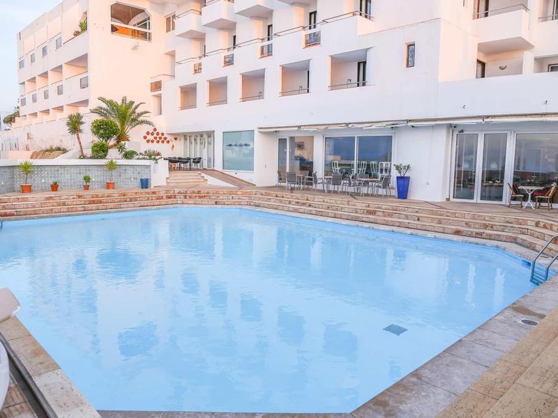 Jasno oświetlony basen hotelowy z leżakami wokół i przylegającym budynkiem.