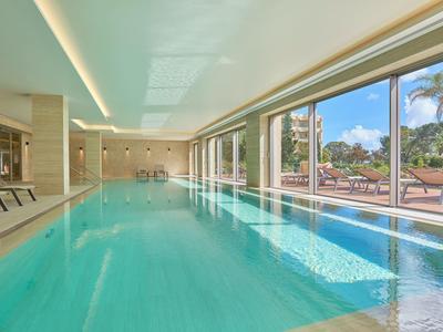 Světle krytý bazén s dřevěnými lehátky a velkými okny s výhledem na zahradu a palmy.