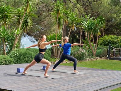 Zwei Frauen praktizieren Yoga im Freien auf einer Holzplattform, umgeben von grünen Pflanzen.