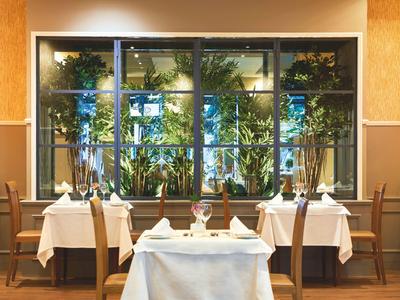 Elegantní restaurace s bílými ubrusy, dřevěným nábytkem a výhledem na zelené rostliny venku.