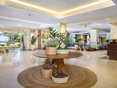 Světlá hotelová hala s kulatými stoly, rostlinami a útulnými posezeními.