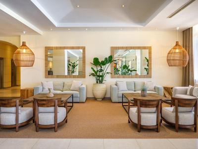 Světlá hotelová lounge se sedačkami, křesly, rostlinami a velkými nástěnnými zrcadly.