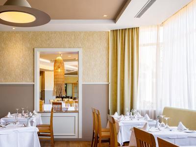 Elegantní restaurace s bílými ubrusy, dřevěnými židlemi a velkými okny se žlutými závěsy.