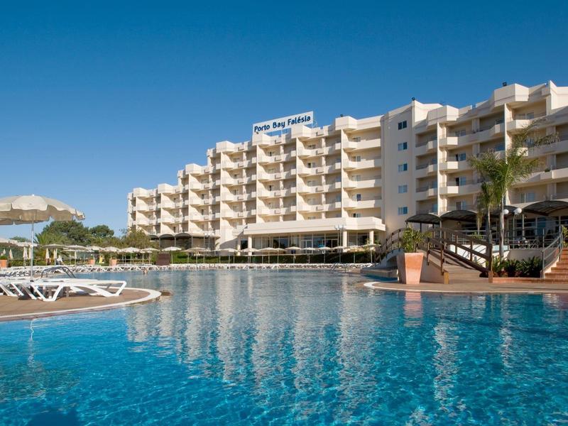 Velká vícepodlažní budova hotelu vedle velkého bazénu pod jasnou modrou oblohou.