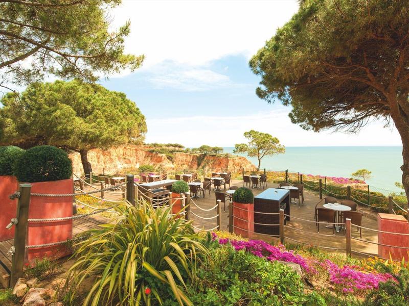 Venkovní terasa s stolem a židlemi obklopená rostlinami s výhledem na moře.