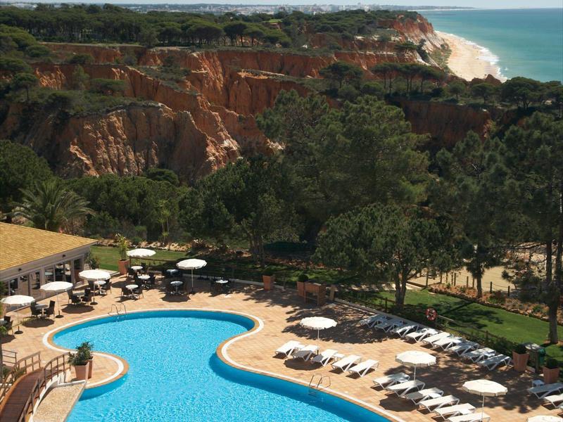 Velký bazén s lehátky a hosty, obklopen stromy a útesem s výhledem na moře.