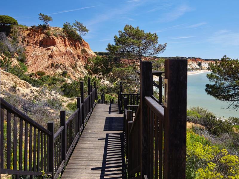 Dřevěná lávka s zábradlím vede podél skalnatého pobřeží s výhledem na moře.